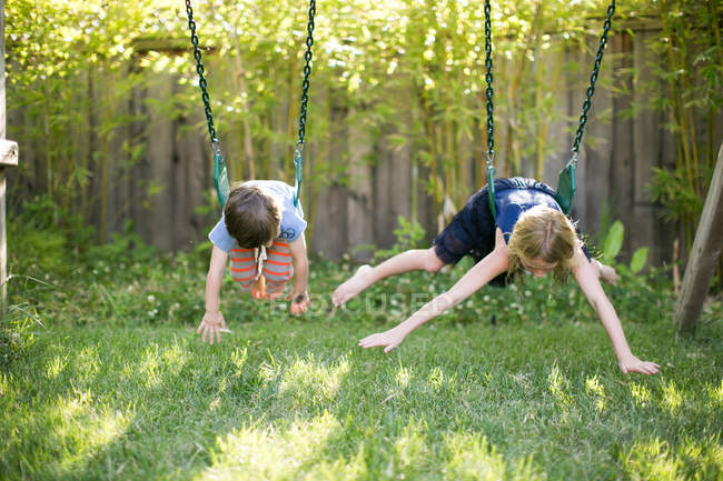 Chico y chica acostados en frentes balanceándose en columpios de jardín - foto de stock