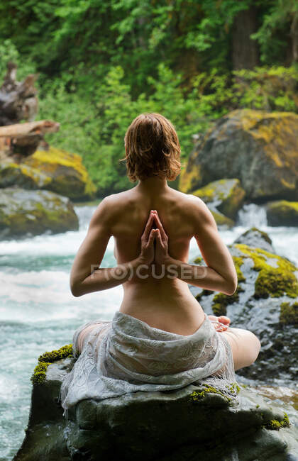 Vista trasera de la mujer en topless meditando en la roca por el agua - foto de stock