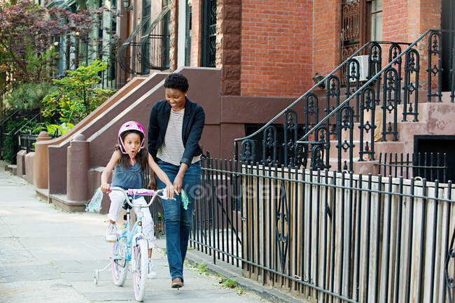 Hija aprendiendo a andar en bicicleta a lo largo de la acera con madre - foto de stock