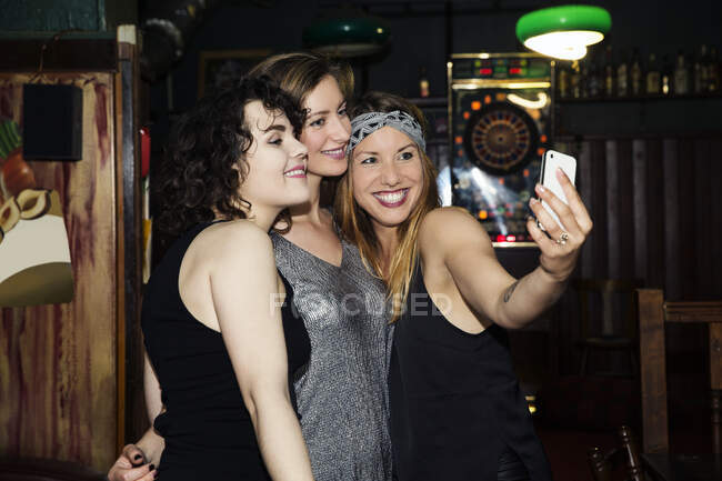 Tres amigas adultas tomando selfie de teléfonos inteligentes en la noche en el bar - foto de stock