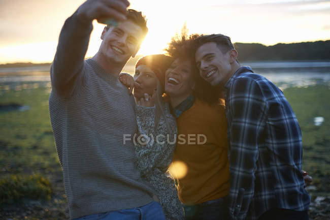 Cuatro adultos jóvenes tomando selfie smartphone al atardecer junto al mar - foto de stock
