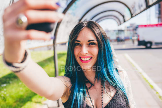 Молода жінка з зануреним пофарбованим блакитним волоссям бере смартфон у міському автобусному притулку — стокове фото