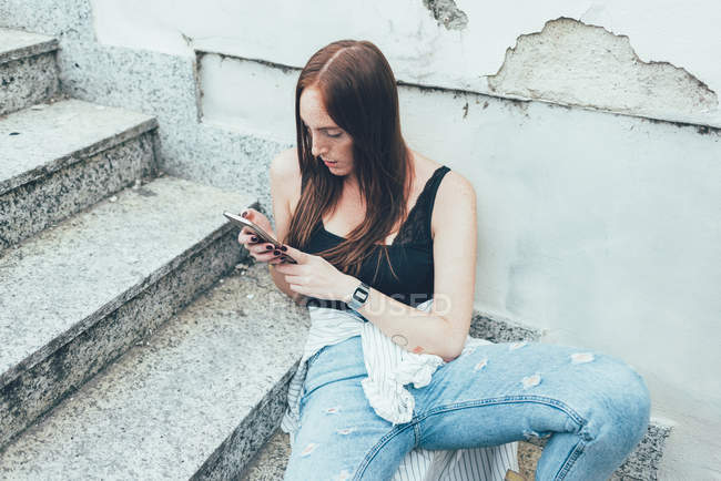 Junge Frau sitzt im Treppenhaus und liest Smartphone-Texte — Stockfoto