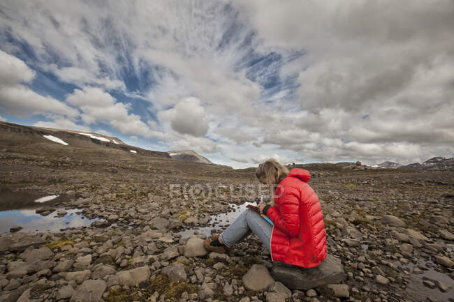 Turista sentada en paisaje rocoso escribiendo en cuaderno, Seyoisfjorour, Islandia - foto de stock
