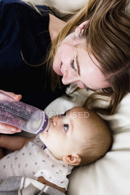 Вид сверху на среднюю взрослую женщину, кормящую дочку бутылочкой на диване — стоковое фото