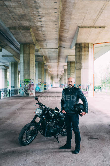 Maduro masculino motociclista em pé por moto sob o viaduto — Fotografia de Stock