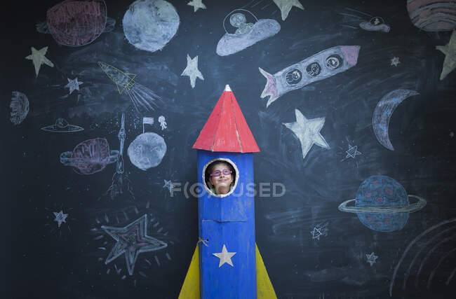 Portrait of girl in handmade space rocket in front of space themed chalk drawings — Stock Photo