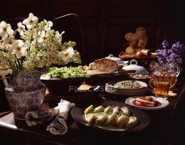 Traditionelle Ausstellung von Lebensmitteln, Blumen in der Vase, Lammbraten, Kartoffeln, Teddybär, Krug Bier, Spaghetti und Kuchen — Stockfoto