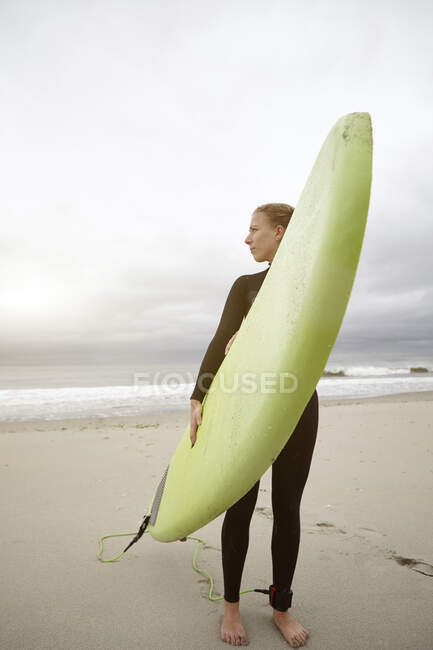 Surfista feminina carregando prancha de surf olhando para trás de Rockaway Beach, Nova York, EUA — Fotografia de Stock