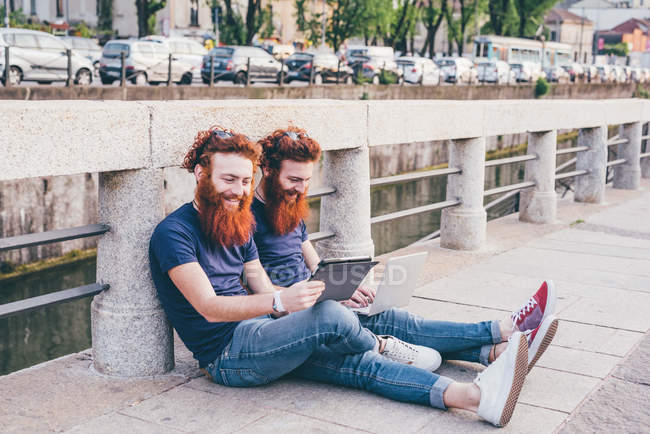 Joven macho gemelos hipster con pelo rojo y barbas navegando tableta digital en puente - foto de stock