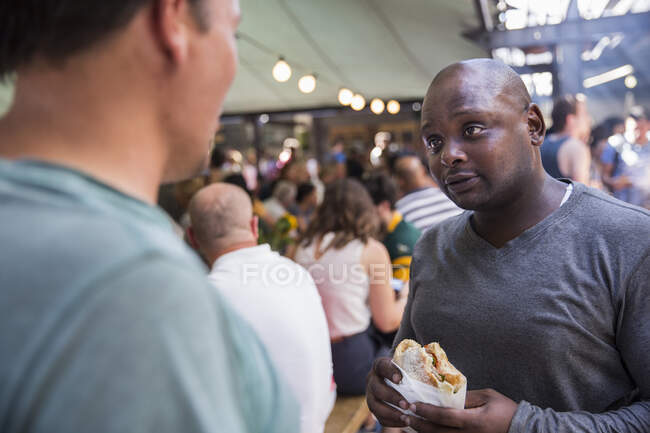 Male customers chatting and eating burgers at cooperative food market stall — Stock Photo