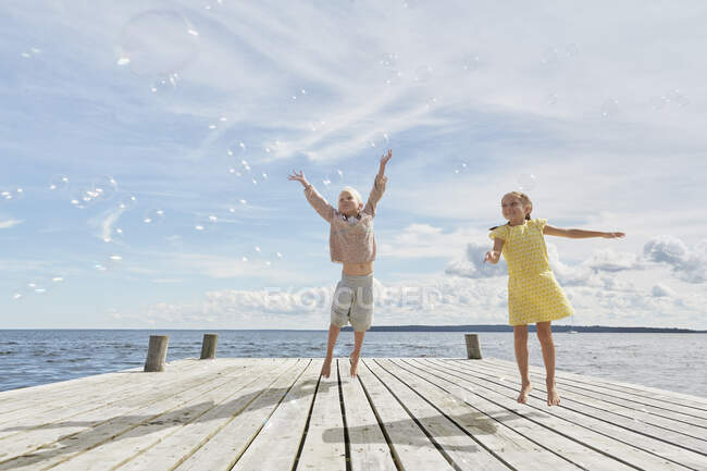 Two young friends on wooden pier, jumping to reach bubbles — Stock Photo
