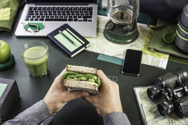 Mans mano in possesso di sandwich a tavola con attrezzature da trekking e laptop — Foto stock