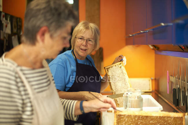 Дві старші жінки-бджолярки обрізають медоносні комбінації на кухні — стокове фото