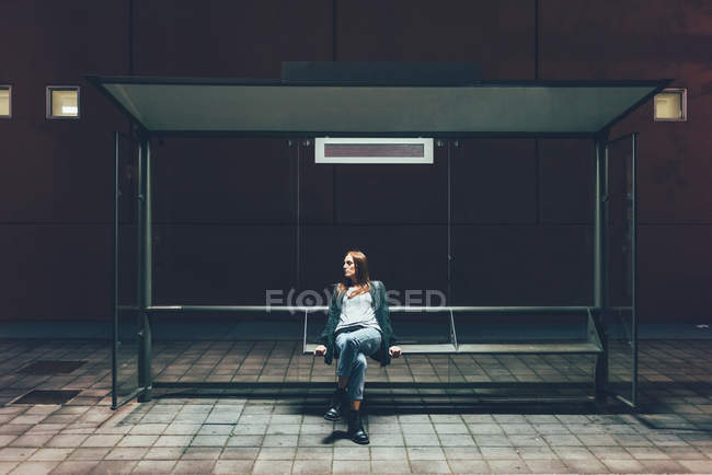Mujer joven sentada en la parada de autobús por la noche - foto de stock