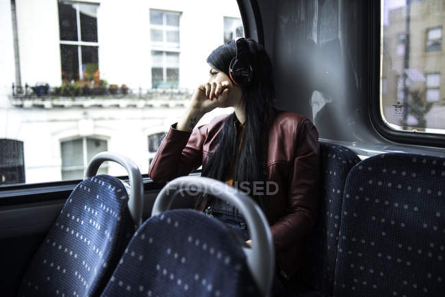 Young woman sitting on bus, wearing headphones, looking out of window — Stock Photo