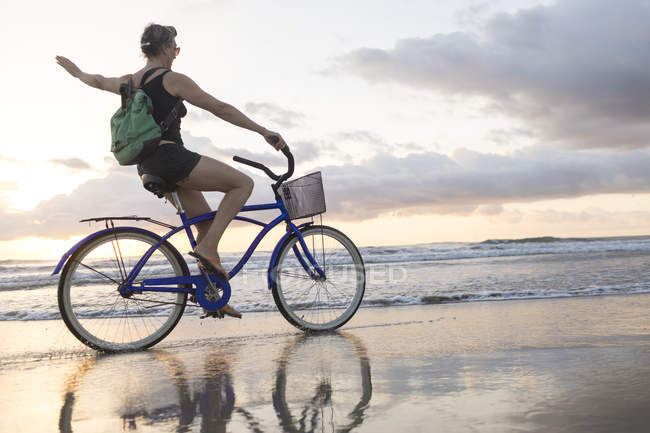 Woman waving while cycling on beach at sunset, Nosara, Guanacaste Province, Costa Rica — Stock Photo