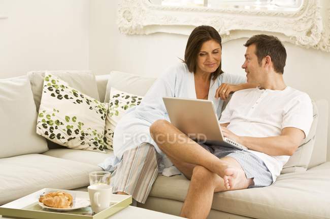 Mature couple on sofa browsing laptop and having breakfast — Stock Photo