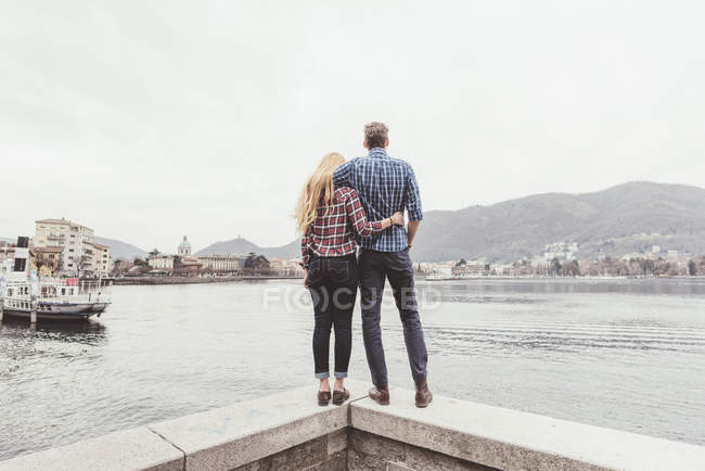 Вид сзади на молодую пару, стоящую на стене гавани и выглядывающую наружу, озеро Комо, Италия — стоковое фото