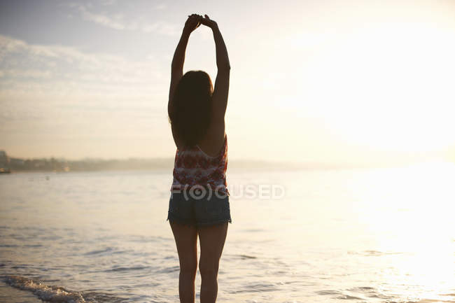 Vista trasera de la mujer de pie junto al mar con los brazos levantados - foto de stock
