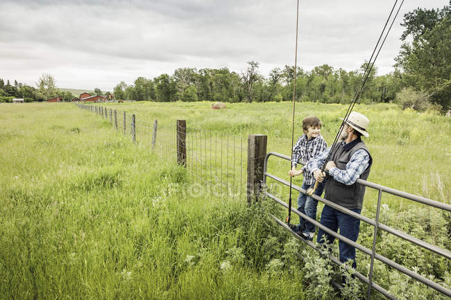 Man and boy holding fishing rods resting on gate in field — Stock Photo