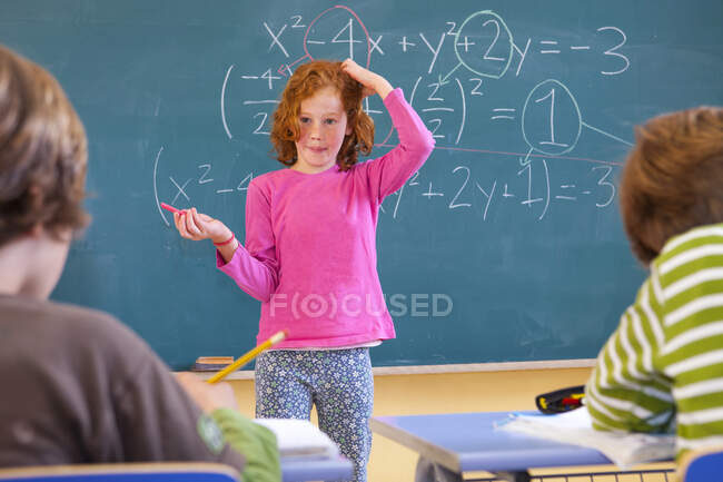 Colegiala primaria arañándose la cabeza en la ecuación en la pizarra del aula - foto de stock