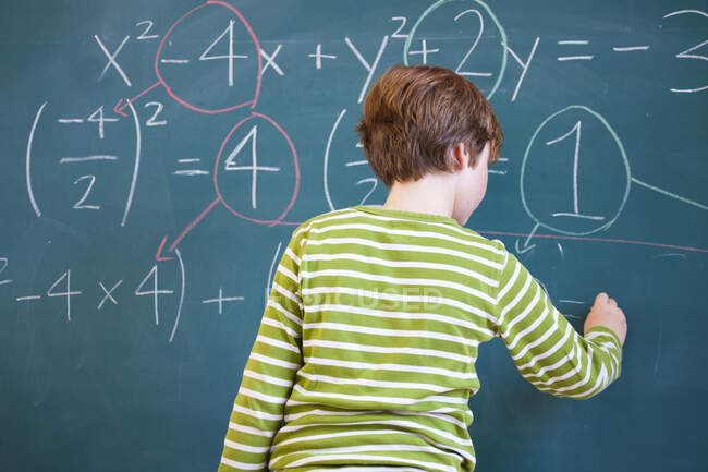 Rear view of primary schoolboy answering equation on classroom blackboard — Stock Photo