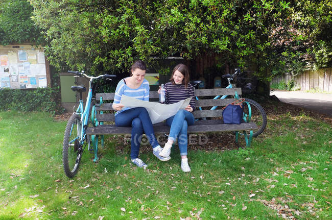 Hermanas adultas jóvenes con bicicletas sentadas en el banco mirando el mapa - foto de stock