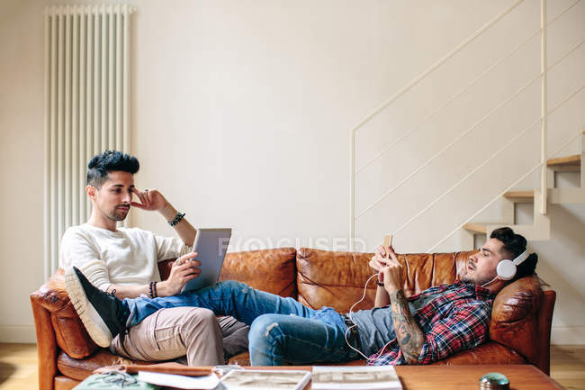 Male Relaxing On Sofa Wearing Headphones Using Smartphone Male Relaxing On Sofa Wearing Headphones Using Smartphone And Laptop Stock Photo