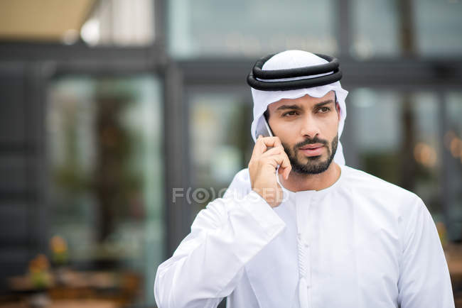 Hombre usando dishdasha caminando por la calle hablando por teléfono inteligente, Dubai, Emiratos Árabes Unidos - foto de stock