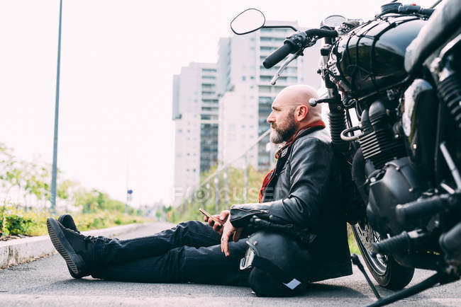 Maduro motociclista do sexo masculino sentado na beira da estrada inclinado na motocicleta com smartphone — Fotografia de Stock