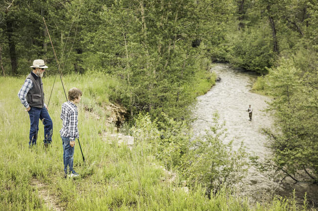 Homme et garçon tenant des cannes à pêche regardant l'homme pêcher dans la rivière — Photo de stock