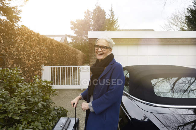 Женщина возле машины на подъездной дорожке, держит чемодан, смотрит в камеру улыбаясь. — стоковое фото