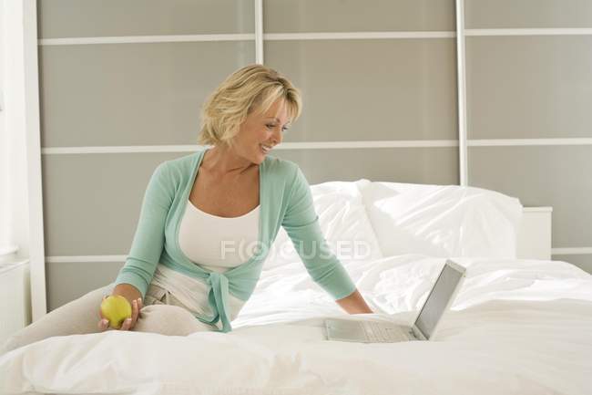 Зріла жінка сидить на ноутбуці перегляду ліжка — стокове фото