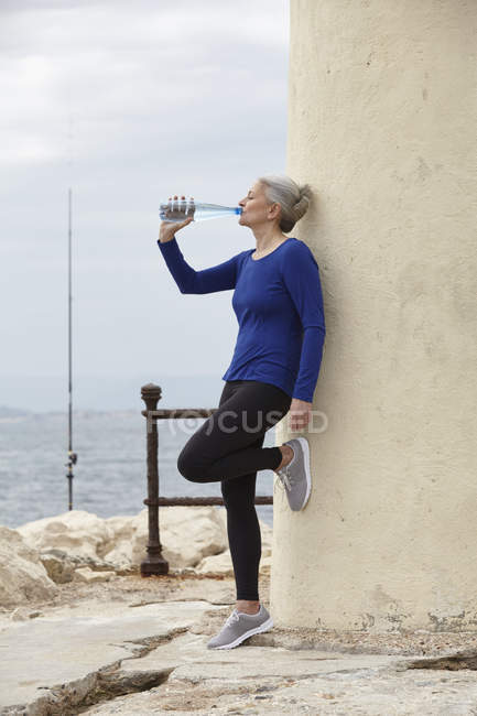 Mature woman outdoors, leaning against wall, drinking from water bottle — Stock Photo