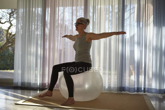 Зрелая женщина балансирует на мяче для упражнений перед дверьми патио — стоковое фото