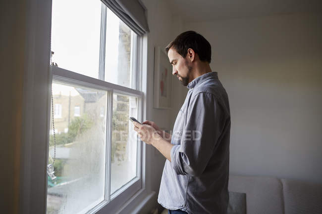 Hombre adulto medio en la ventana de la sala de estar leyendo textos de teléfonos inteligentes - foto de stock