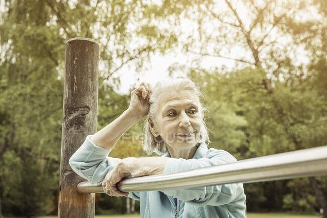 Mulher sênior no parque encostado à barra de metal olhando para longe sorrindo — Fotografia de Stock