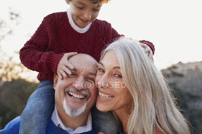 Бабушка и дедушка с внуком на плечах смотрят в камеру улыбаясь — стоковое фото