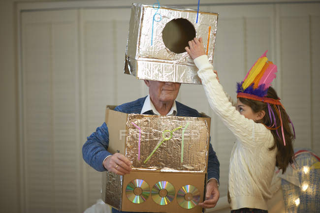 Girl in feather headdress putting robot costume onto grandfather — Stock Photo