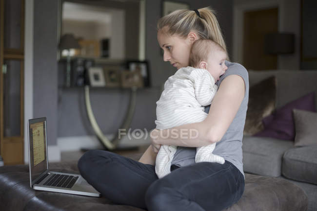 Mother holding baby boy whilst using laptop — Stock Photo