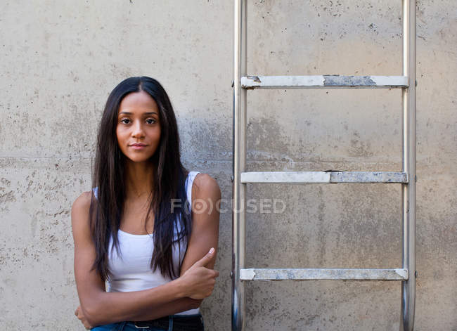 Mulher encostada à parede de concreto ao lado da escada, braços cruzados olhando para a câmera — Fotografia de Stock