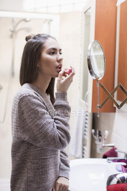 Young woman, looking in bathroom mirror, applying lipstick — Stock Photo