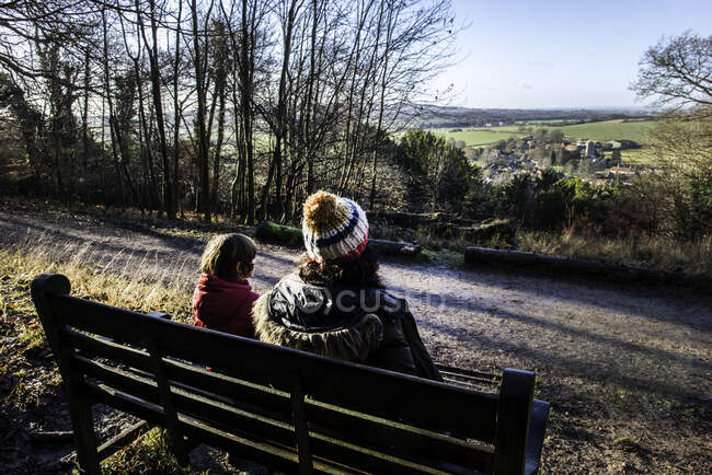Madre e hijo sentados en el banco en el entorno rural, mirando a la vista, vista trasera - foto de stock