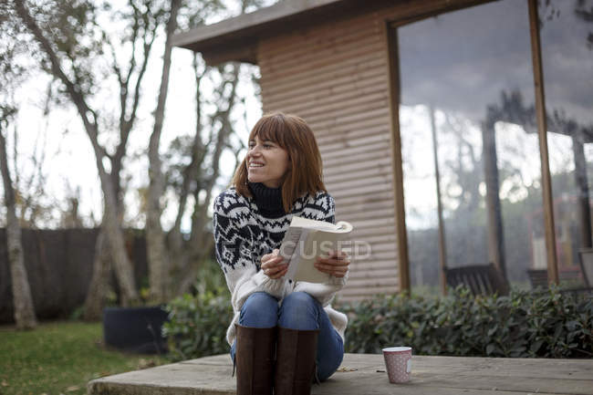 Жінка сидить на столі для пікніка тримає книгу, дивлячись геть усміхнений — стокове фото
