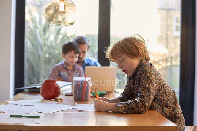 Мальчик раскрашивает за обеденным столом, пока отец использует ноутбук с братом — стоковое фото