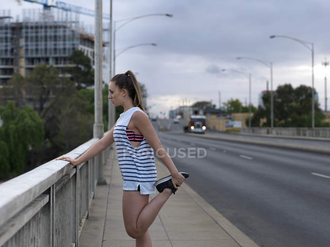 Jeune coureuse étirant les jambes sur un pont routier à l'aube — Photo de stock