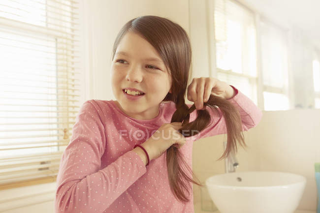 Девушка заплетает длинные каштановые волосы в ванной комнате — стоковое фото