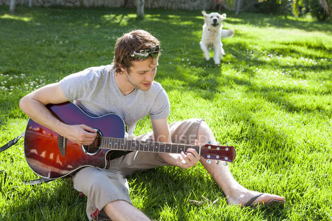 Молодой человек сидит в саду и играет на акустической гитаре — стоковое фото