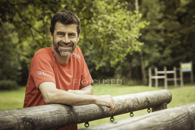 Hombre maduro apoyado en el equipo del patio de recreo mirando a la cámara sonriendo - foto de stock